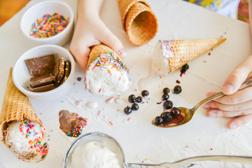 The child Makes his own Ice cream: Top View, Children's Hands Holding a cone of ice Cream On the table Chocolate, Spoon, Topping, Berries, Sprinkles, Jam.