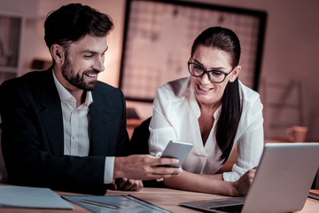 Fototapeta premium I will show you. Satisfied beardful pleasant man sitting in the cabinet near a woman smiling and showing her his cellphone.