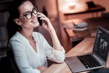 Important call. Occupied pleasant bespectacled woman sitting in the office by her desktop having phone conversation and using the laptop.