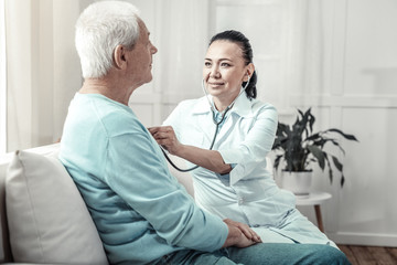 Fototapeta premium Medical procedure. Young skilled pleasant nurse sitting in the white room on the sofa listening to the her patient lungs and using her stethoscope.