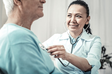 Breathe deeply. Mature joyful pretty doctor sitting near her patient using the stethoscope and smiling.