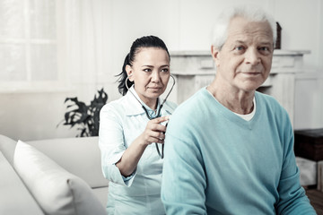 Skilled nurse. Cute serious reliable nurse sitting in the room behind the man listening to the patients body and concentrated on him.