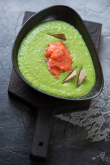 Bowl of green peas cream-soup with salmon on a black wooden serving board, grey concrete background, studio shot