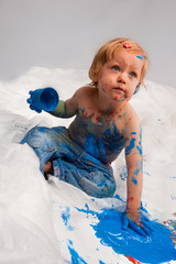 zwei Jahre altes Mädchen im Studio vor weißem Hintergrund, schmiert blaue Farbe auf den Boden und ist selbst mit Farbe beschmiert