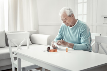 Struggle with the illness. Lonely serious aged man sitting in the bright room holding pack of tablets and overlooking pills.