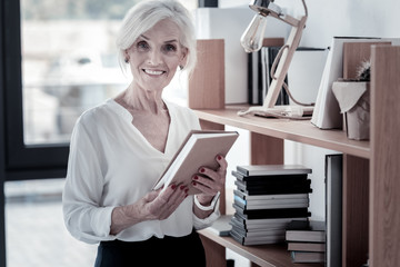 Follow me. Positive delighted mature woman keeping smile on her face and holding notebook while looking straight at camera