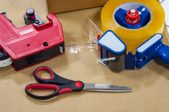 Packing tape dispenser, scissors and label gun on the cardboard box.