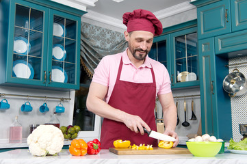 Man in hat preparing vegetables. Guy with knife cutting orange pepper on table. Cook dressed in...