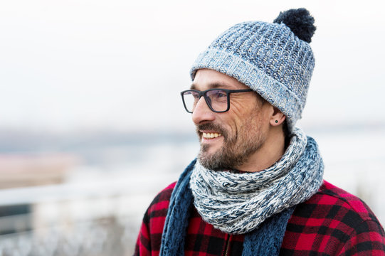 Profile portrait of young smiling man in glasses and red jacket. Winter knitted clothes for urban man. Profile of happy bearded guy in glasses and knitted scarf. Buddy in hat with ball.