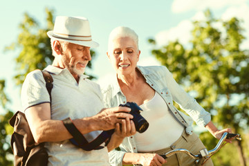 What we have. Positive nice aged man holding a camera and showing photos on it while standing together with his wife