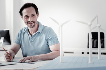 Be positive. Handsome bearded architect keeping smile on his face and turning head while leaning arms on the table