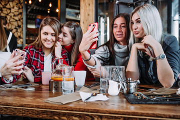 Four friends sitting by the table in cafe and making selfie on smartphone