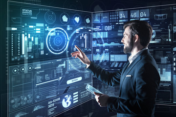 Satisfying results. Cheerful enthusiastic young programmer feeling happy while standing in his modern convenient office and pointing to the diagrams on a transparent screen