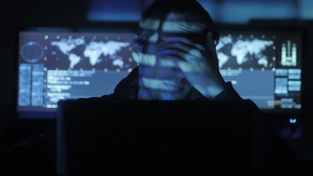 Man geek hacker in hood overworking at computer and suffers from a headache in cyber security center filled with display screens.
