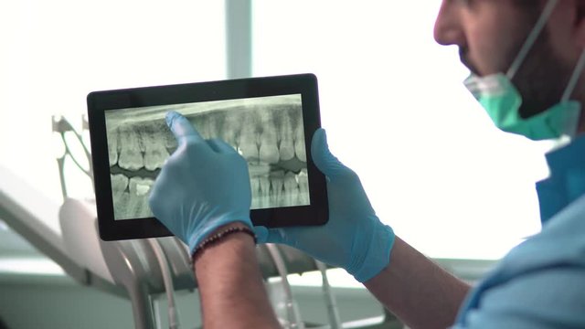 Intelligent dentist showing teeth x-ray on tablet, well-built man in green mask and blue gloves explaining particular case, sitting before dental chair in light huge office