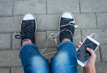 Teen girl in jeans and sneakers sitting outdoors, using a smartphone and headphones in the hands, close up, white smart phone.