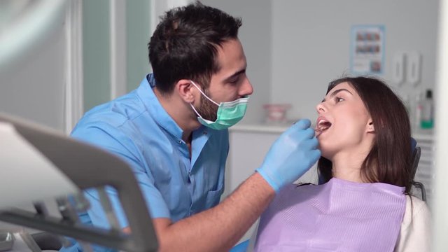 Dentist using mouth mirror, obedient patient opening the mouth, doctor explaining the problem while examining teeth, indoor shot in well-equipped dental office