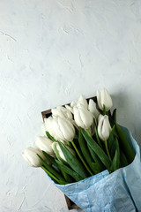A bouquet of white tulips in blue wrapping paper in wooden box on a white concrete background. Top view. Flat lay. Postcard for Easter, Mother's Day and Spring Holidays.
