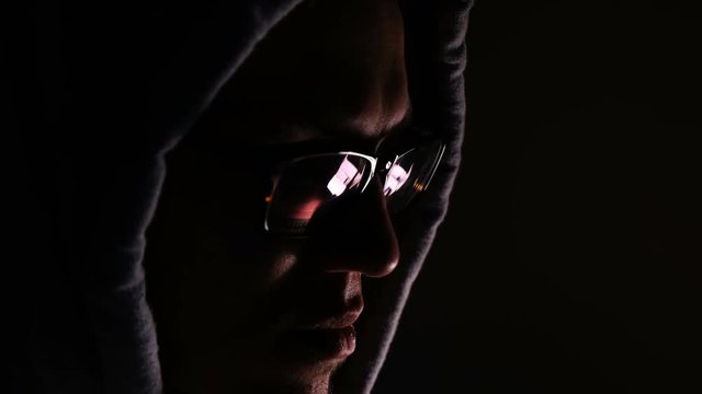 Hacker in glasses and a hood works at a computer in the dark, a reflection in glasses close-up, 4k