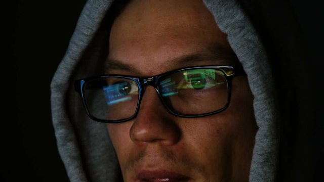 Hacker in glasses and a hood works behind a computer in the dark, the program code is reflected in glasses close-up, 4k