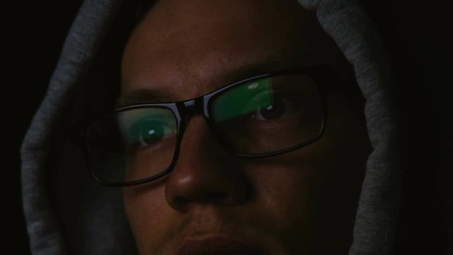 Programmer with glasses and a hood works at a computer in the dark, a reflection in glasses close-up, 4k