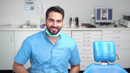 Smiling young dentist using tablet, well-built man in blue uniform and gloves doing short research while waiting for a patient near dentist chair in light huge office - Powered by Adobe