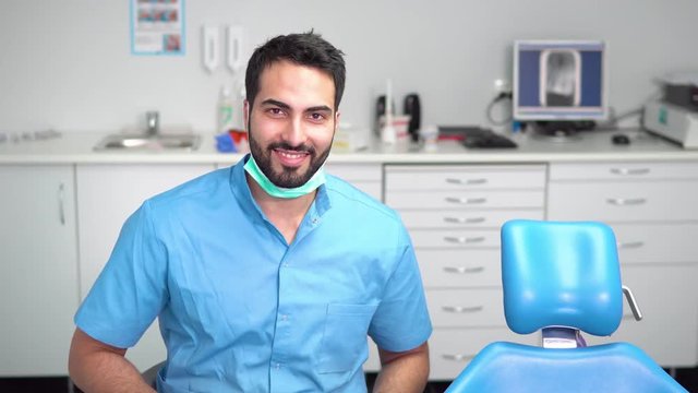 Smiling young dentist using tablet, well-built man in blue uniform and gloves doing short research while waiting for a patient near dentist chair in light huge office