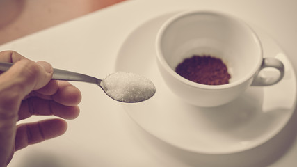 Closeup of hand pouring sugar from spoon in cup with coffee, image with warm vintage toning