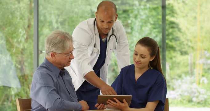 4K Senior man with friendly doctor & nurse, looking at tablet & discussing healthcare