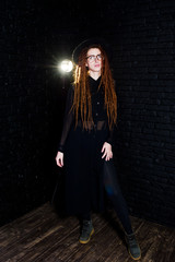 Studio shoot of girl in black with dreads, at glasses and hat on brick background.