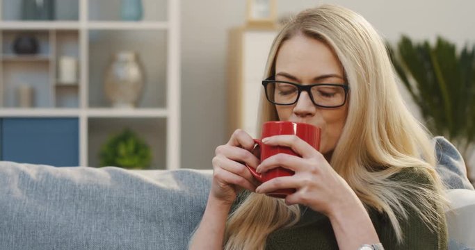 Portrait shot of the pretty blonde woman in glasses drinking a tea from a red cup while sitting on the sofa in the living room. Indoors