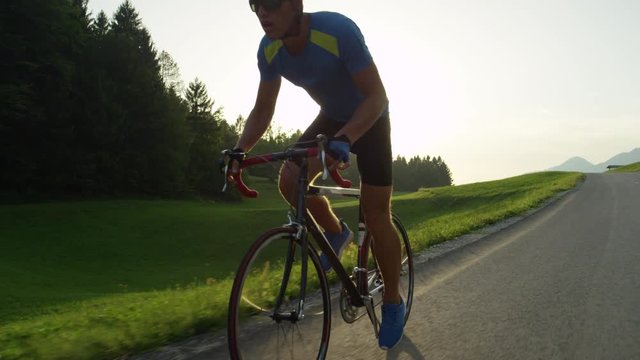 SLOW MOTION, LENS FLARE: Male biker pedals his road bicycle past the camera on a sunny evening. Young athlete riding his bike during race in the spring countryside. Pro cyclist easily overtakes camera