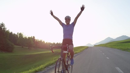 LENS FLARE: Professional biker raising his arms victoriously after winning race. Cheerful biking athlete celebrates victory in quiet countryside. Pro cyclist cheerfully rides road bike with no hands.
