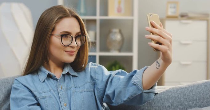Close up of the young pretty woman in the glasses making a selfie on the smartphone on the couch in the cozy room. Inside