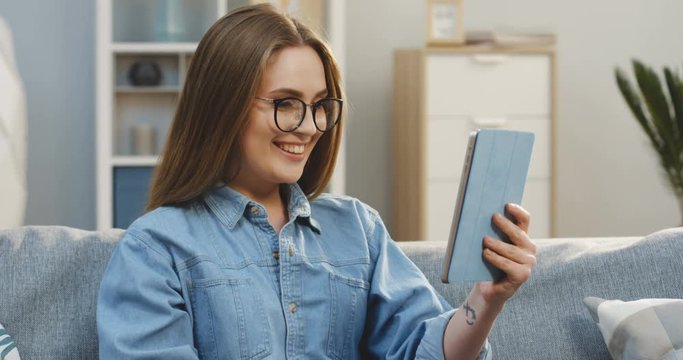 Portrait of the atractive young woman in the jeans shirt and glasses having video chatting on the tablet computer in the cozy living room. Indoor