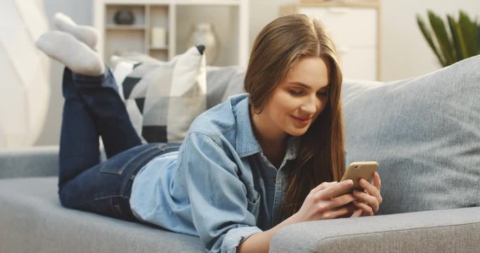 Pretty young woman in the jeans clothes taping on the smartphone and smiling while lying on the couch at home. Indoor