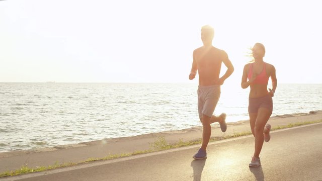 SLOW MOTION, COPY SPACE, SUN FLARE: Sporty woman and man on holiday running near the beautiful ocean on a perfect afternoon in summer. Active couple running alone along the tranquil sunlit ocean road.