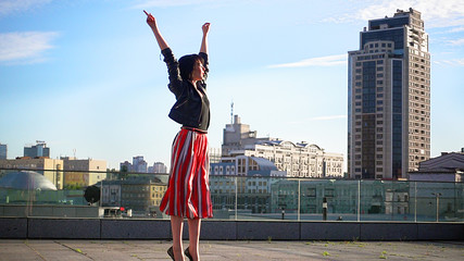 Fototapeta na wymiar Beautiful brunette woman is dancing feeling happy outdoors. City life urban young happy people behavior. Sunset cityscape clear sky shot. The woman wears long striped pleated skirt.