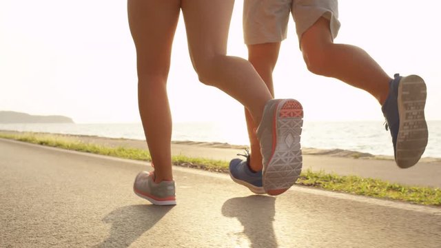 SLOW MOTION, LOW ANGLE, SUN FLARE: Young athletic woman and man wearing training shoes run along sunny ocean road. Unrecognizable Caucasian girlfriend and boyfriend jog on a sunny day at the beach.