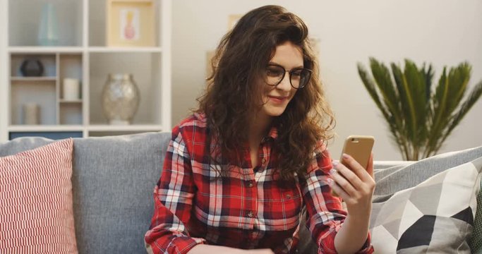 Portrait of the atractive young woman in the plaid shirt and glasses having video chatting on the smartphone in the cozy living room. Indoor