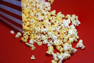 Heap of popcorn from red and white paper box on the red floor. It is the corn of a variety with hard kernels that swell up and burst open with a pop when heated.