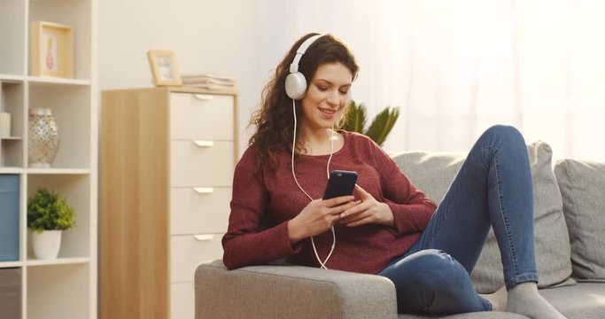 Portrait of the charming young woman with curly hair in the white headphones listening to the music on the smartphone and smiling while sitting on the sofa at home. Indoor