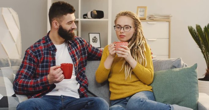 Attractive hipsters woman and man drinking a tea or coffee while sitting on the sofa at home and talking. Portrait shot. Indoors