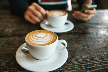 Two cups of coffee on a wooden table, a man holding a phone in his hand and going to call. Waiting for a meeting. Another person is late for the meeting.