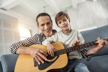 Spending time together. Handsome cheerful fair-haired boy learning to play the guitar while sitting on the couch and his father smiling