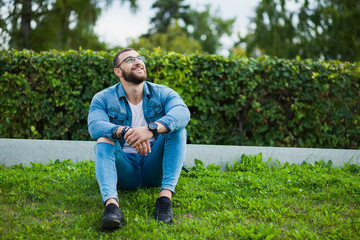 Naklejka na ściany i meble Young strong muscular guy sit on grass and relax. Male fitness model in jeans wear denim casual look, hipster man with beard and glasses.