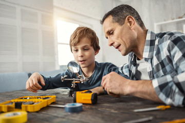 Teaching my son. Handsome concentrated dark-haired man showing instruments to his son while sitting at the table and his son holding a magnifying glass
