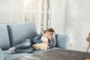Daydream. Cute little fair-haired boy hugging his favourite nice toy and lying on the comfy sofa...
