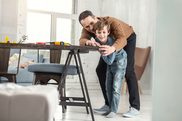 Devoted father. Nice joyful little fair-haired boy holding a measuring tape and measuring the table and his daddy helping help