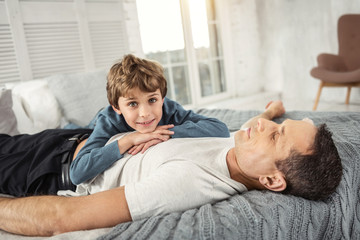 My best father. Nice delighted fair-haired boy smiling and his daddy while lying on his back on the sofa and they relaxing together